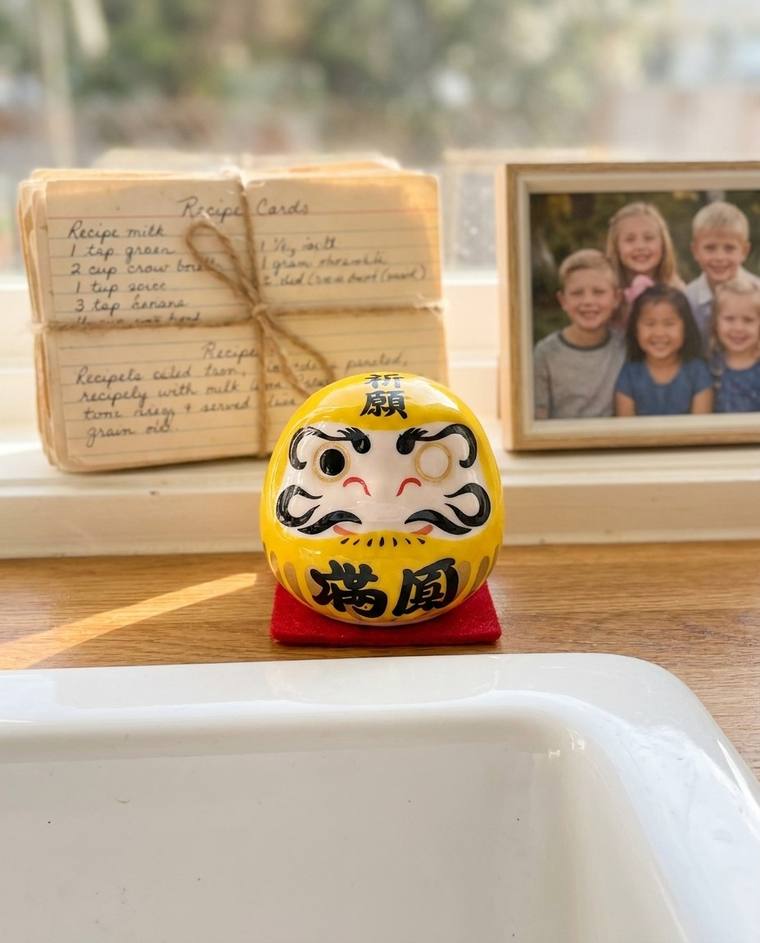 A one-eyed yellow Daruma on the counter beside a phone charger