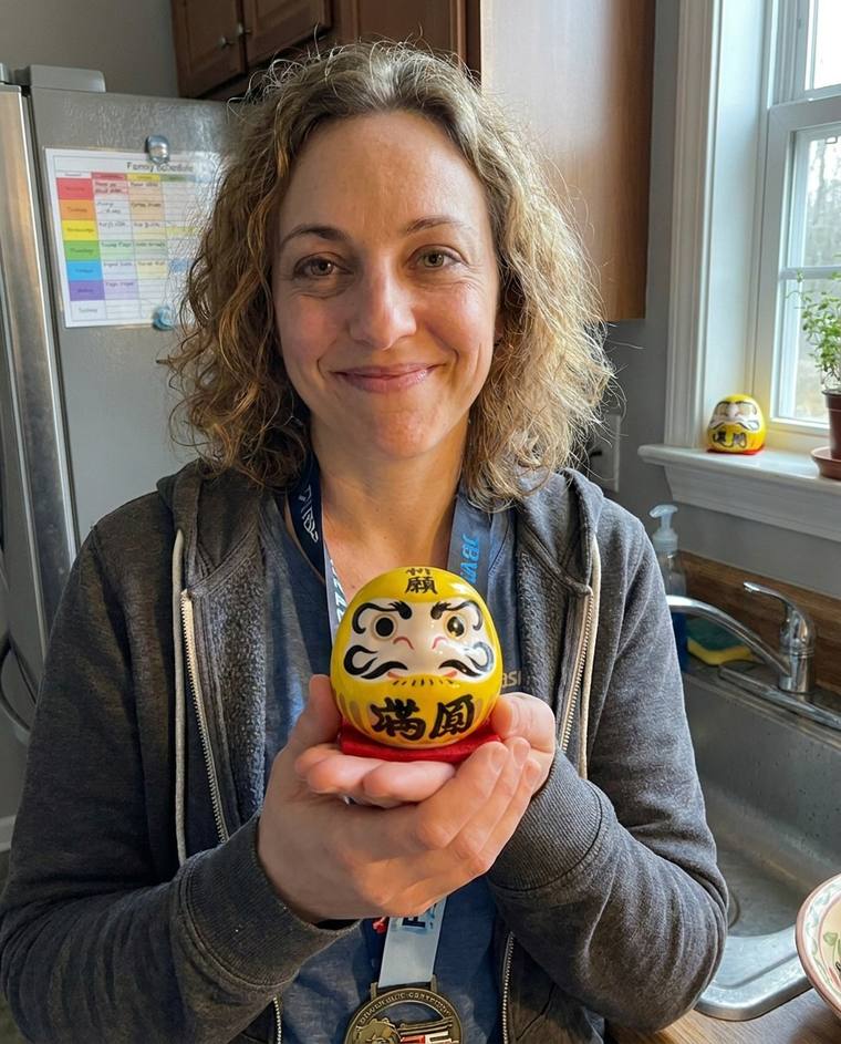 Mariah painting the second eye of the yellow Daruma at the kitchen sink, still wearing a race bib