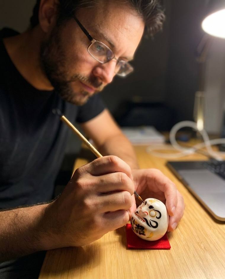 Tyler filling in the first eye of a white Daruma at his desk before dawn
